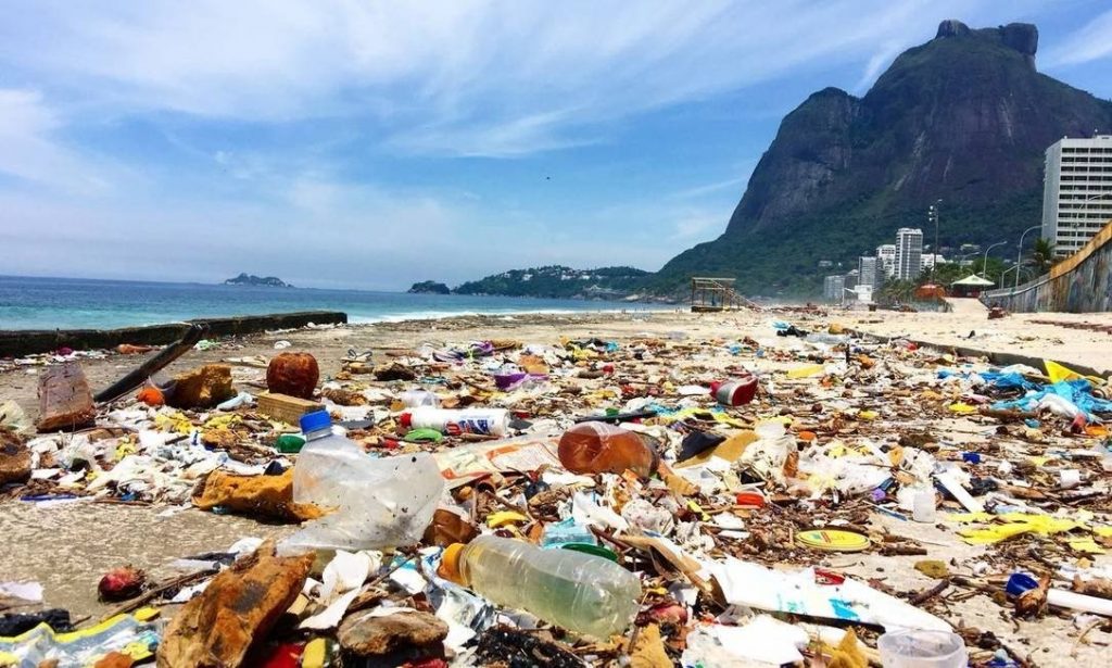 Foto de uma praia com as areias cobertas com diversos tipos de lixo, principalmente plásticos. Mar e Morro Dois Irmãos ao fundo.