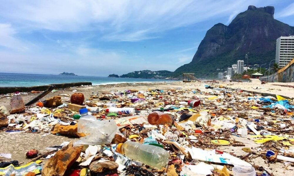 Foto de uma praia com as areias cobertas com diversos tipos de lixo, principalmente plásticos. Baia de Guanabara e Morro Dois Irmãos ao fundo.